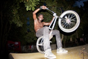 A topless man in black jeans sits on a BMX bike with his front wheel raised in the air