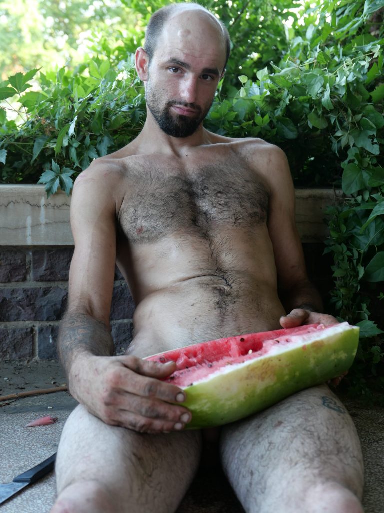 A balding white man with a beard poses with a watermelon poised in front of his private parts