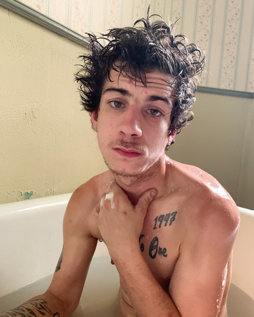 A naked tattooed white man with messy hair stares down the lens of a camera while sat in a bath tub