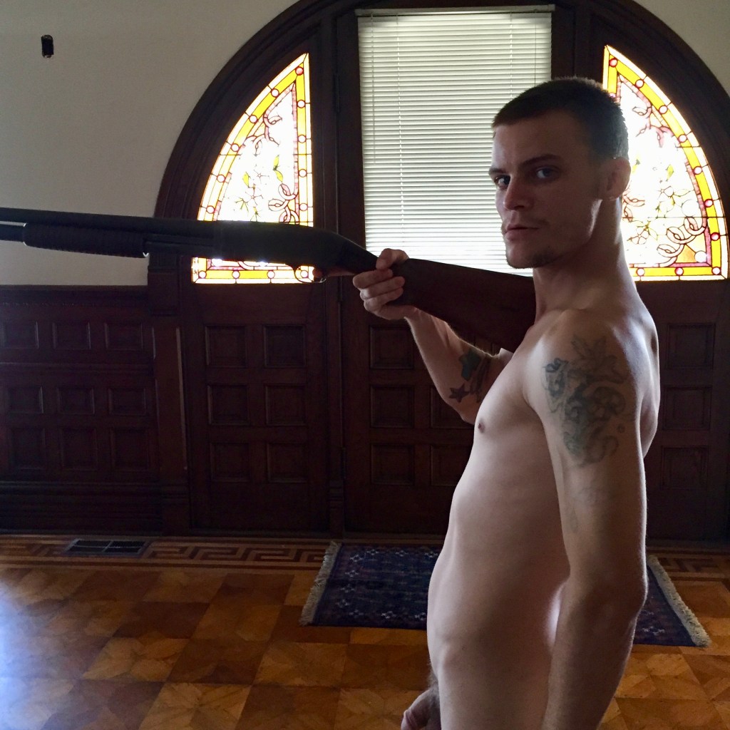 A white man poses naked with a shotgun while looking down the lens of a camera