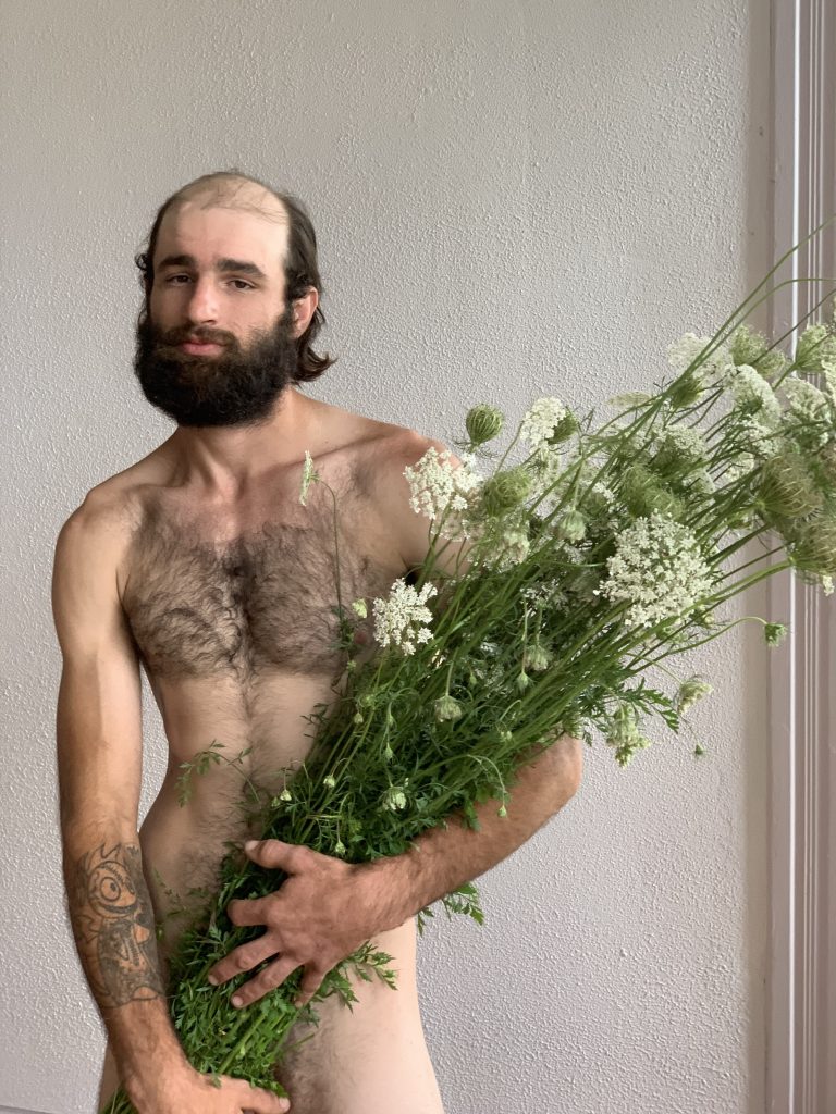 A balding white man with a combover and bushy beard poses naked with flowers covering his private parts