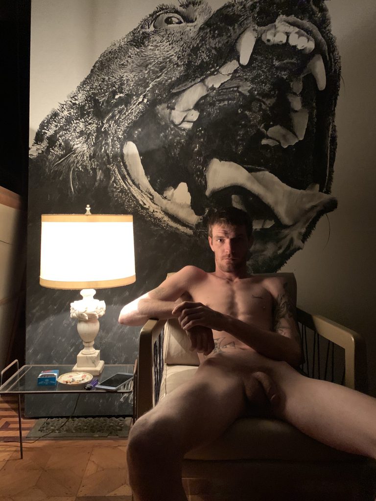 A naked white man reclines on a chair next to a lamp and in front of a large photograph of a dog's jaws